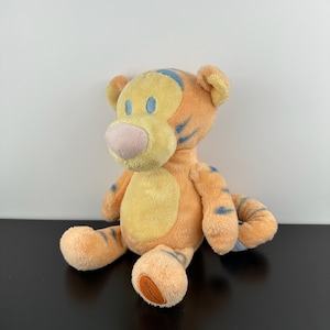 May include: A plush tiger toy featuring soft orange and yellow fur with light blue stripes. The character has a light pink nose, light blue oval eyes, and a circular orange patch on the bottom of one foot. The stuffed animal is in a seated position.