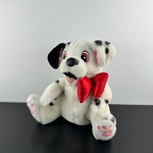 May include: A plush dalmatian dog toy with white fur, black spots, and a large red satin bow around its neck. The stuffed animal features black ears, orange and white eyes, a black nose, and pink paw pads on its feet while sitting upright.
