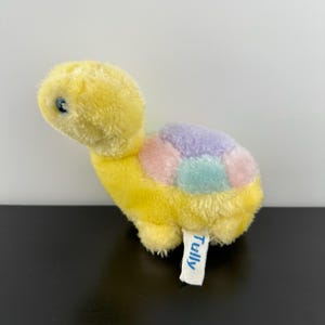 May include: A soft yellow turtle toy with a shell in pastel colours of pink, lilac, and turquoise. The toy has a small blue eye and a tag that reads "Tully".