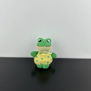 May include: A small plush frog toy with a green head and limbs, featuring a yellow torso decorated with green polka dots. The frog has large black and white eyes, a stitched red mouth, and a light yellow ribbon tied around its neck.