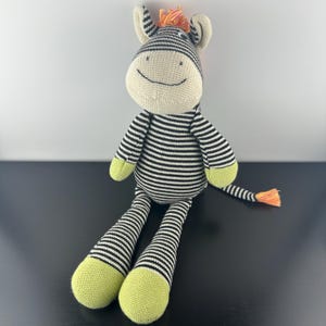 Może przedstawiać: Pluszowa zabawka w kształcie zebry z czarno-białym pasiastym korpusem, nogami i ramionami. Zebra ma kremową twarz, uszy i stopy. Grzywa i ogon są pomarańczowe. Zabawka siedzi na czarnej powierzchni.