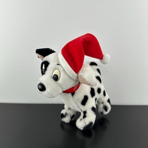 May include: A plush dalmatian puppy toy with black spots on white fur, wearing a bright red Santa hat with a white trim and a small white pom-pom at the tip. The stuffed animal has a red collar around its neck and large, expressive eyes.