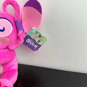 Op de afbeelding: Een roze Stitch-knuffel met grote oren en een label met het Stitch-logo en afbeeldingen van watermeloen, een drankje en een citroen. Het pluche speelgoed heeft een levendige roze kleur.