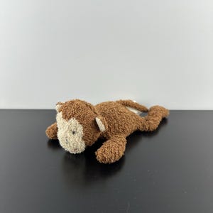 Puede incluir: Un peluche de mono hecho de tela marrón texturizada con una cara de color crema. El animal de peluche está en posición tumbada con las extremidades extendidas, presentando orejas pequeñas, ojos bordados y una nariz en su zona facial clara.