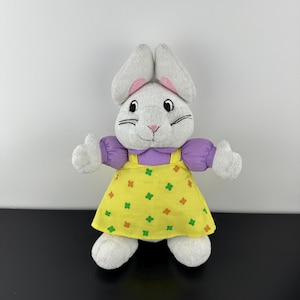 Op de afbeelding: Een witte pluche konijnenknuffel met lange oren met roze accenten aan de binnenkant. Het konijn draagt een paars shirt met korte mouwen onder een gele tuinbroekjurk met een herhalend patroon van kleine groene en oranje klaverblaadjes.