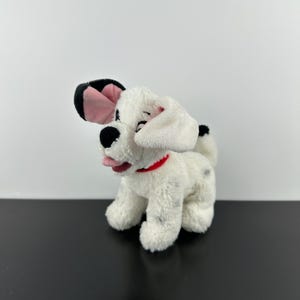 May include: A plush dalmatian puppy toy with white fur and black spots. The stuffed animal features one black ear and one white ear with a pink inner lining, a black nose, a small pink tongue, and a thin red collar around its neck.