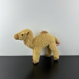 Op de afbeelding: Een pluche kameel in tinten bruin en beige. Het knuffeldier heeft een harige textuur, kleine zwarte ogen en een kleine bult. De kameel staat op vier poten, met een label aan de achterkant.