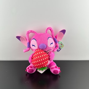Op de afbeelding: Een roze pluche speelgoedfiguur dat een rode aardbei met witte stippen vasthoudt. Het speelgoed heeft grote oren, paarse accenten en gesloten ogen. Een klein label is aan het speelgoed bevestigd.