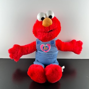 Può includere: Peluche rosso Elmo con una salopette blu con un cuore rosa e un pesce rosso all'interno. Elmo ha grandi occhi bianchi e un naso arancione brillante.