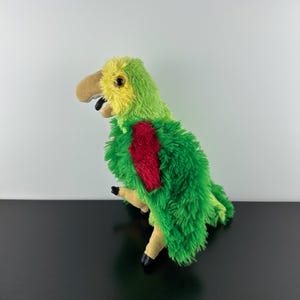 May include: A plush parrot toy with green, yellow, red, and tan coloring. The toy has a large beak, black feet, and a red patch on its chest. The parrot is made of soft, fuzzy material and is designed to resemble a real parrot.