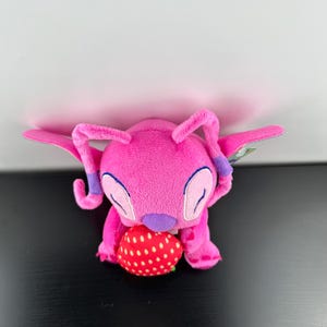 Op de afbeelding: Een roze pluche speelgoedfiguur met grote oren en vleugels, die een rode aardbei met witte stippen vasthoudt. Het speelgoed heeft paarse accenten en staat tegen een witte en zwarte achtergrond.