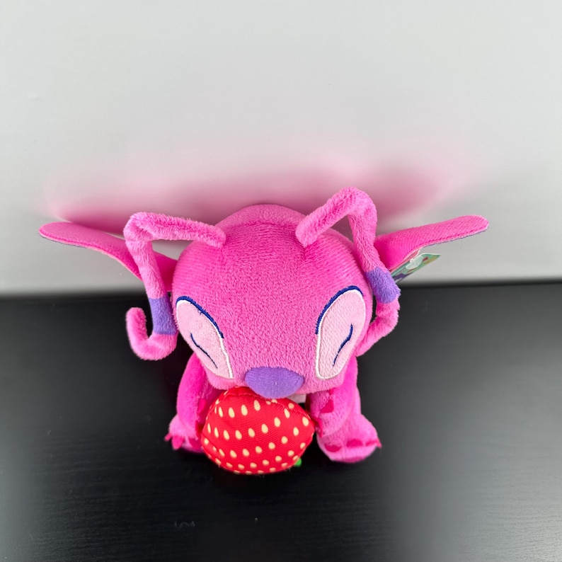 Op de afbeelding: Een roze pluche speelgoedfiguur met grote oren en vleugels, die een rode aardbei met witte stippen vasthoudt. Het speelgoed heeft paarse accenten en staat tegen een witte en zwarte achtergrond.