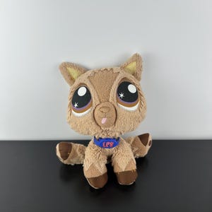 Puede incluir: Un peluche de llama con ojos grandes y expresivos y un collar azul. La llama es principalmente marrón con detalles en marrón oscuro en las patas y las orejas. El collar tiene las letras "LPS" en rojo.