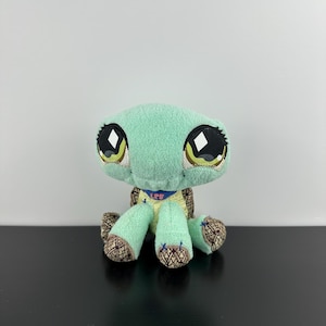 May include: A plush turtle toy with mint green fabric, large black eyes featuring white diamond-shaped highlights, and a blue collar with the pink text LPS. The shell and feet have a brown and tan cross-hatched pattern on the fabric surface.