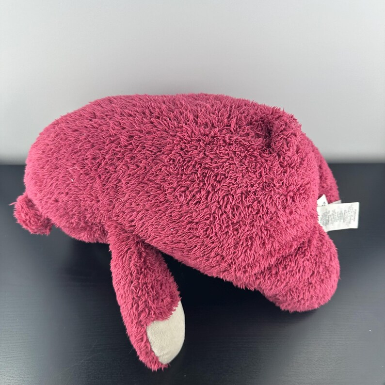May include: A plush, burgundy-colored bear-shaped pillow. The pillow has a soft, fuzzy texture and features a light gray paw. A white tag is attached to the pillow.