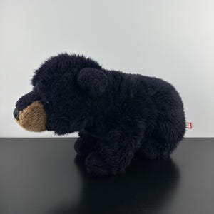 May include: A black plush bear toy with a brown snout and a small red tag. The bear is on a black surface against a white background. The bear's fur looks soft and fluffy.