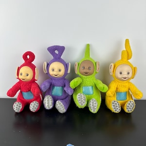 May include: Four plush Teletubbies dolls in red, purple, green, and yellow. Each doll has a smiling face, a screen on its belly, and a unique antenna shape. The dolls are sitting on a black surface against a white background.