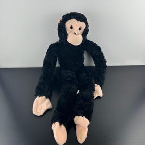 May include: A plush chimpanzee toy featuring long, black, fuzzy fur covering its body and limbs, with a light beige face, ears, hands, and feet. The stuffed animal has dark, round eyes and a stitched nose and mouth, with long arms and legs.