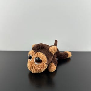 May include: A small plush monkey toy featuring dark brown fur on its body and lighter tan fur on its face, ears, and paws. The stuffed animal has large, round, dark brown eyes and a long, slender tail extending from its back in a lying position.