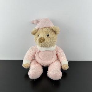 Op de afbeelding: Een pluche teddybeer met een roze outfit en bijpassende nachtmuts. De beer heeft een lichtbruin gezicht, oren en neus, met zwarte ogen. De roze outfit heeft een witte kraag en manchetten.