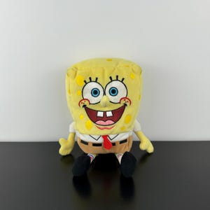 May include: A plush yellow SpongeBob SquarePants toy with embroidered features, including large blue eyes, a wide smile, and a red tie. The toy is wearing a white shirt, brown trousers, and black shoes. The toy is sitting.