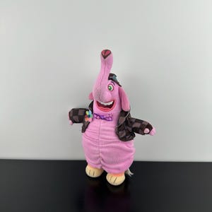 May include: A pink plush elephant character with a long trunk, green eyes, and a wide smile. It wears a brown and black checkered jacket, a purple polka-dot bowtie, and a small multi-colored flower accessory on its chest. The plush has light tan feet.