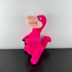 Può includere: Un peluche di dinosauro rosa acceso con un becco beige chiaro e una bocca cucita in nero. Il dinosauro ha piccoli artigli neri ed è ambientato su uno sfondo bianco.