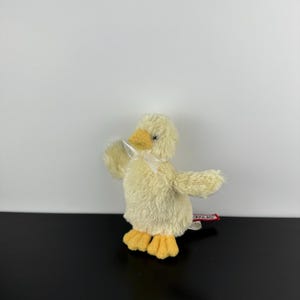 May include: A plush yellow duck toy with a yellow beak and feet, and a white ribbon around its neck. The duck has outstretched wings and a tag attached to its body. The toy is on a black surface.