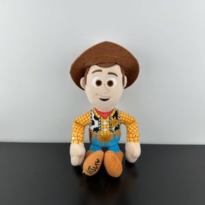 May include: A soft toy of the character Woody from the film Toy Story. Woody is wearing a brown cowboy hat, a yellow plaid shirt, blue jeans, and brown boots. The word "Andy" is written on the bottom of the boots.