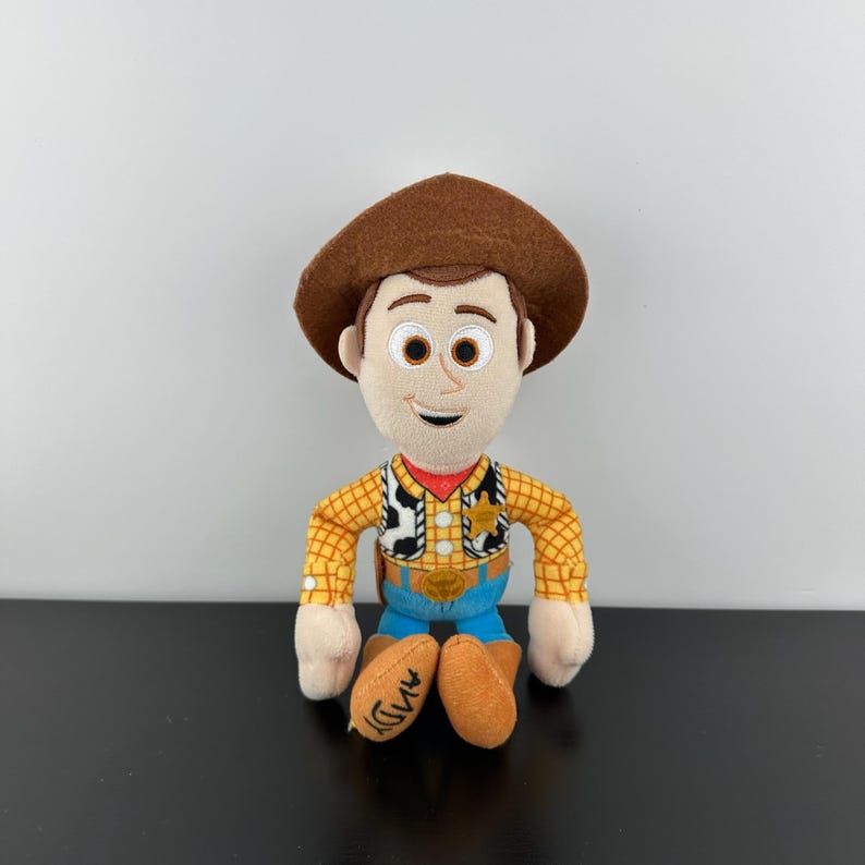 May include: A soft toy of the character Woody from the film Toy Story. Woody is wearing a brown cowboy hat, a yellow plaid shirt, blue jeans, and brown boots. The word "Andy" is written on the bottom of the boots.