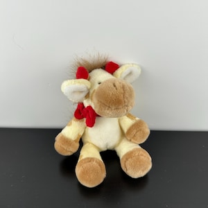May include: A plush cow toy with cream and light brown fur, featuring a brown snout, small red horns, and a bright red bow tied around its neck. The stuffed animal has a tuft of brown hair on its head and soft, rounded ears, sitting in an upright position.