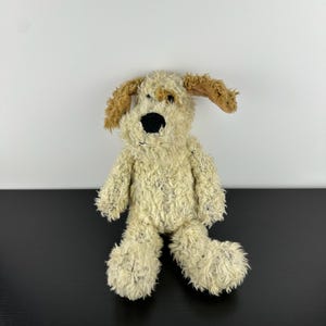 May include: A plush dog toy with a cream-colored body and brown ears. The dog has a black nose and a friendly expression. The toy is made of soft, textured material and is designed to be cuddly.