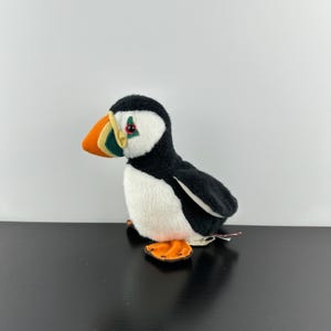 May include: A plush puffin toy with a black back, wings, and head, a white belly, and orange feet. The bird has a large, colorful beak with orange, yellow, and green stripes. It has a red eye and is set against a white background.