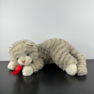 May include: A plush gray tabby cat toy with white paws and a red ribbon. The stuffed animal is lying down on a black surface. The cat has a pink nose and is made of soft, textured material.