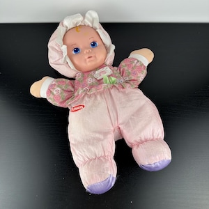 Può includere: Una bambola vintage Playskool con un corpo rosa tenue e un top con stampa floreale. La bambola ha gli occhi blu, un viso dipinto e un cappuccio bianco. I piedi della bambola sono viola. Il logo Playskool è visibile sul corpo della bambola.