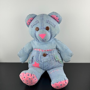 Puede incluir: Un oso de peluche hecho de tela vaquera azul claro. Presenta ojos de botón marrones, una nariz triangular de color rosa brillante y detalles de tela rosa con patrones de estrellas azules en las orejas y los pies. El oso lleva pantalones cortos vaqueros.