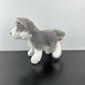 May include: A soft toy husky dog, featuring grey and white fur. The toy dog has a black nose and is posed on all fours. The fur is fluffy, and a small red tag is attached to its rear.