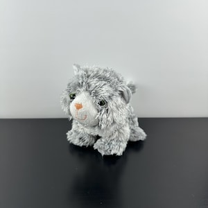 Puede incluir: Un juguete de peluche gris con forma de gato, con ojos verdes y nariz rosa. El peluche tiene un aspecto esponjoso y texturizado y está colocado sobre una superficie negra sobre un fondo blanco. El gato está sentado.