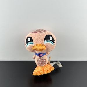 Puede incluir: Un juguete de peluche de pato con ojos grandes y expresivos y un pico naranja cosido. El pato tiene un cuerpo rosa con detalles estampados y patas naranjas. Un collar azul alrededor de su cuello dice "LPS".