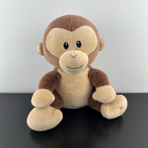 May include: A plush monkey toy featuring soft brown and tan fur. The monkey has a rounded head with a light tan face, dark embroidered eyes, and a stitched mouth. Its arms and legs are brown with tan hands and feet, and it has a tan belly.