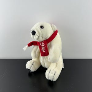 Puede incluir: Oso polar de peluche blanco con una bufanda roja con el logotipo de Coca-Cola.