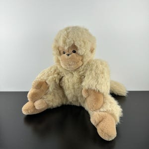 May include: A plush toy monkey with light brown fur and a tan face, hands, and feet. The stuffed animal is sitting with its legs spread and arms outstretched. The monkey has a small tail and dark eyes.