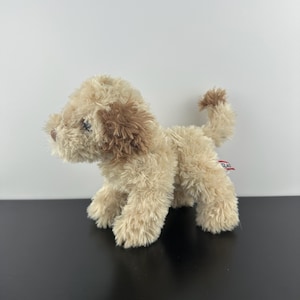 Puede incluir: Un perro de peluche con pelaje desgreñado de color beige claro y orejas de color marrón oscuro. El animal de peluche está de pie en posición de perfil, con una pequeña nariz marrón, ojos oscuros y una etiqueta roja y blanca en la parte trasera.