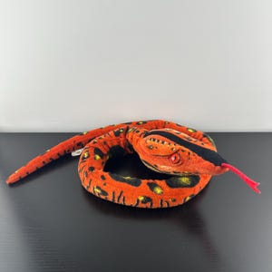 May include: A plush snake toy in shades of orange, black, and yellow. The snake has a black stripe down its head and a red tongue. The toy is coiled and appears to be made of soft fabric.