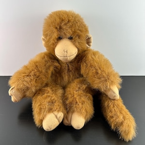 May include: A plush orangutan toy with brown fur, tan face, hands, and feet. The stuffed animal is sitting with its arms and legs spread out. The toy has a long tail.