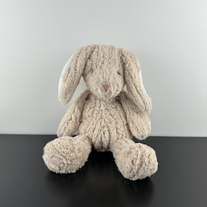 May include: A plush stuffed animal in the shape of a rabbit with long, floppy ears. The toy is covered in textured, beige-colored faux fur and features a small, triangular pink nose. The rabbit is in a seated position with its arms and legs extended.