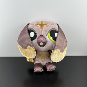 Puede incluir: Un perro de peluche con pelaje marrón y dorado, ojos grandes verdes y negros y una nariz rosa. El juguete está sentado sobre una superficie negra.