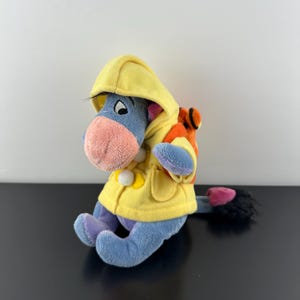 Może przedstawiać: Pluszowa zabawka Eeyore w żółtym płaszczu przeciwdeszczowym z kapturem z białymi pomponami i małym pomarańczowo-żółtym plecakiem. Osiołek jest niebieski z różowym nosem i fioletowymi stopami.