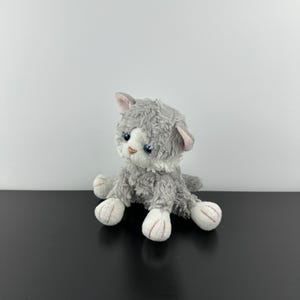 May include: A plush gray kitten with white paws and a white chest sits on a black surface. The stuffed animal has blue eyes, pink inner ears, and a fluffy texture. The kitten is a soft toy.