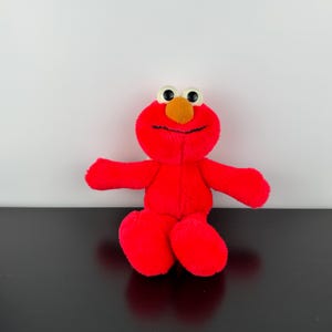 Può includere: Un peluche rosso di Elmo con un naso giallo, un sorriso nero e grandi occhi rotondi. Il personaggio di Sesame Street ha le braccia tese e un aspetto morbido e coccoloso. Il giocattolo è ambientato su uno sfondo bianco e nero.